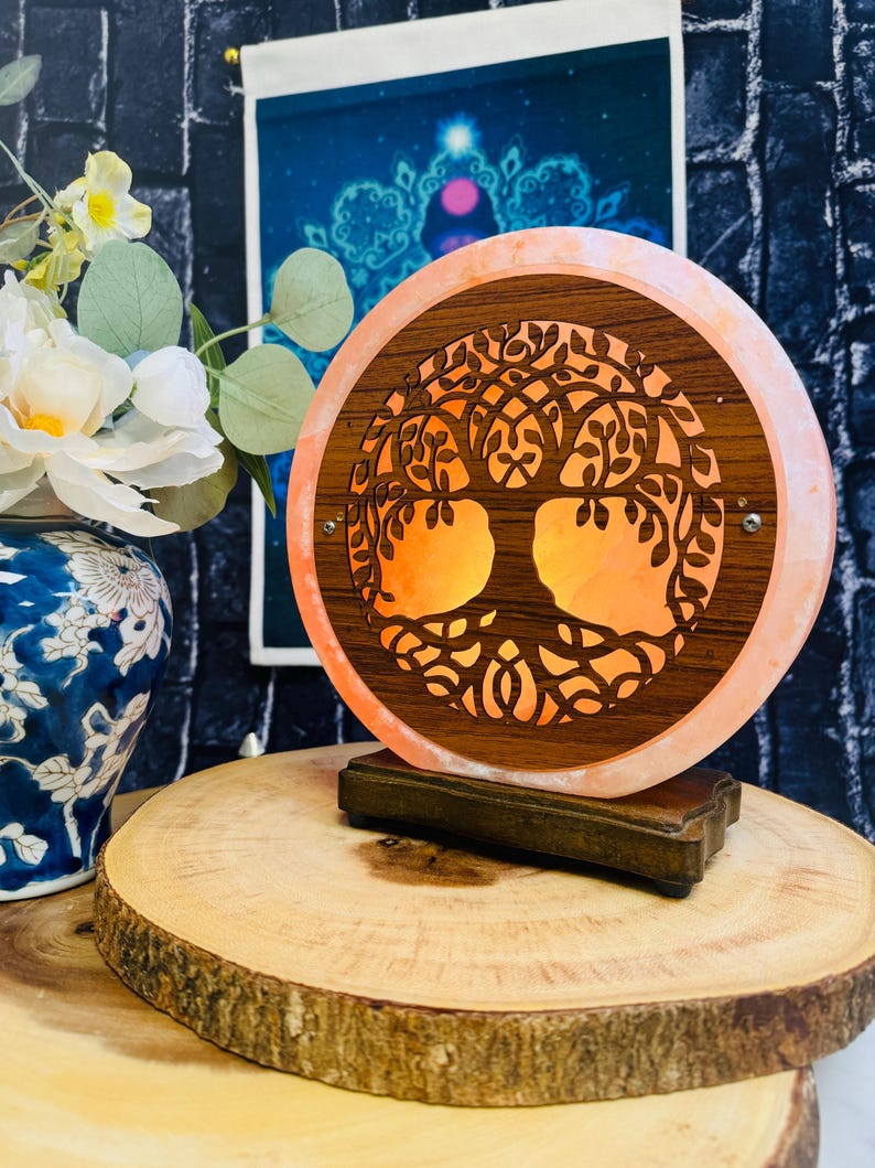 May include: A round Himalayan salt lamp with a wooden tree of life design. The lamp is lit and glows orange. The lamp is on a wooden base and is sitting on a wooden surface.