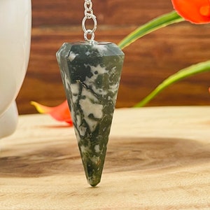 May include: A green and white agate crystal pendulum with a silver chain. The pendulum is pointed and has a faceted shape.
