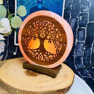 May include: A pink Himalayan salt lamp with a wooden tree of life design. The lamp is illuminated and casts a warm glow. The lamp is on a wooden base.
