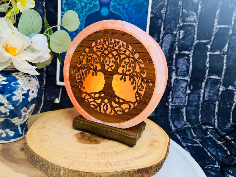 May include: A pink Himalayan salt lamp with a wooden tree of life design. The lamp is illuminated and casts a warm glow. The lamp is on a wooden base.
