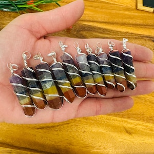 May include: A collection of seven chakra stone pendants, each wrapped in silver wire. The stones are layered in the traditional chakra colors: purple, indigo, blue, green, yellow, orange, and red.