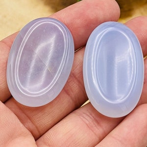 May include: Two oval-shaped, light blue, polished stones with a smooth, shiny surface. The stones have a subtle, iridescent sheen.