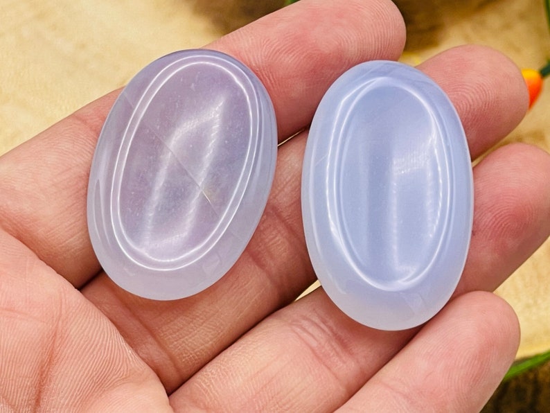 May include: Two oval-shaped, light blue, polished stones with a smooth, shiny surface. The stones have a subtle, iridescent sheen.