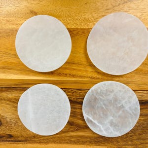 May include: Four white selenite charging plates on a wooden surface. The plates are round and have a smooth, polished finish.