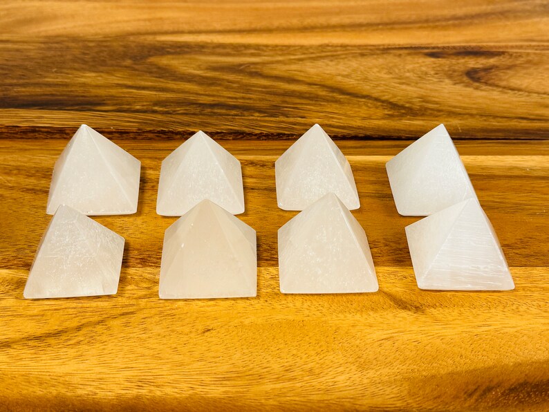 Puede incluir: Ocho pir&aacute;mides de cristal de selenita blanca dispuestas en dos filas de cuatro sobre una superficie de madera.