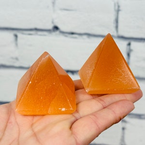 May include: Two orange calcite crystal pyramids on a hand. The pyramids are smooth and have a slight shine.