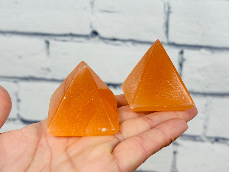 May include: Two orange calcite crystal pyramids on a hand. The pyramids are smooth and have a slight shine.