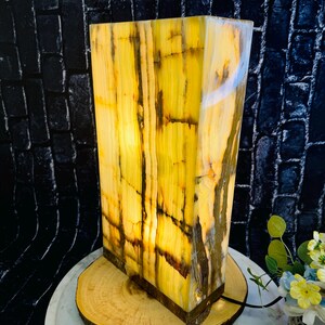 May include: A rectangular onyx lamp with a warm yellow and brown color pattern. The lamp is lit and has a warm glow. It is sitting on a wooden base.