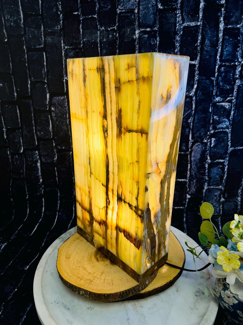 May include: A rectangular onyx lamp with a warm yellow and brown color pattern. The lamp is lit and has a warm glow. It is sitting on a wooden base.