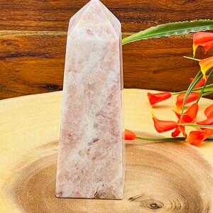 Peut inclure: Une pointe de cristal rose et blanche, &eacute;galement appel&eacute;e ob&eacute;lisque de cristal, est pr&eacute;sent&eacute;e sur une surface en bois. Le cristal a une finition lisse et polie et une pointe pointue.