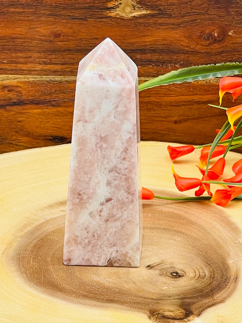 Peut inclure: Une pointe de cristal rose et blanche, &eacute;galement appel&eacute;e ob&eacute;lisque de cristal, est pr&eacute;sent&eacute;e sur une surface en bois. Le cristal a une finition lisse et polie et une pointe pointue.