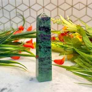 May include: A polished crystal tower featuring a hexagonal prism shape with a pointed top. The stone displays a mottled pattern of vibrant green, deep purple, and black mineral inclusions throughout its smooth, vertical, and reflective surface.