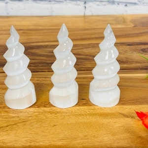 May include: Three white selenite crystal towers with a spiral design. The towers are arranged in a row on a wooden surface.
