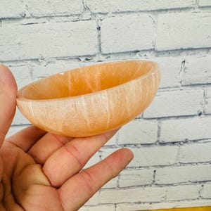 May include: A small, light orange bowl made of selenite crystal. The bowl has a smooth, polished surface and is ideal for holding small items such as jewellery or crystals.