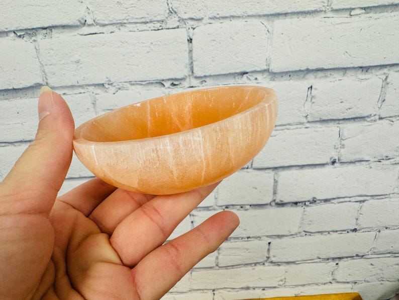 May include: A small, light orange bowl made of selenite crystal. The bowl has a smooth, polished surface and is ideal for holding small items such as jewellery or crystals.