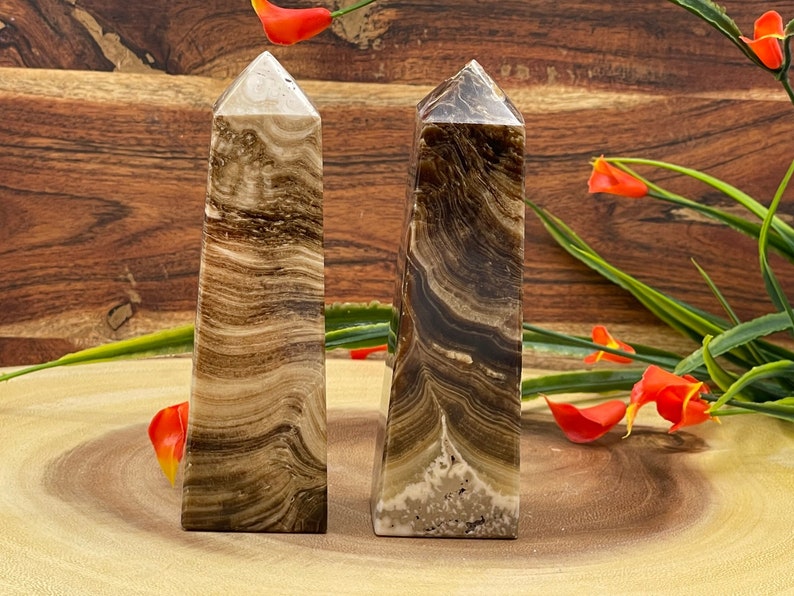 May include: Two brown and cream-colored crystal obelisks with a layered, banded pattern. The obelisks have a rectangular base and taper to a pointed top. The background features a wooden surface and decorative flowers.