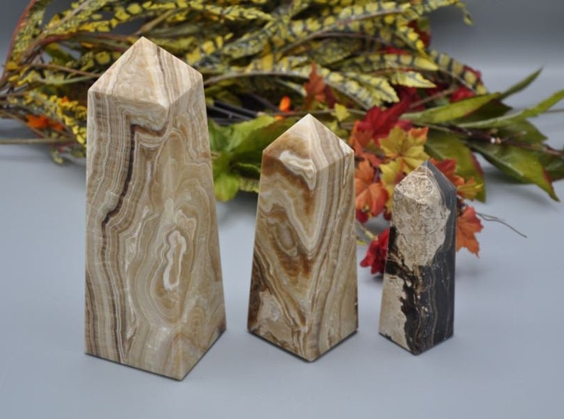 May include: Three stone obelisks with a geometric shape. The obelisks have a beige and brown marbled pattern. The obelisks are of varying heights. The background includes fall foliage with red, orange, and green leaves.