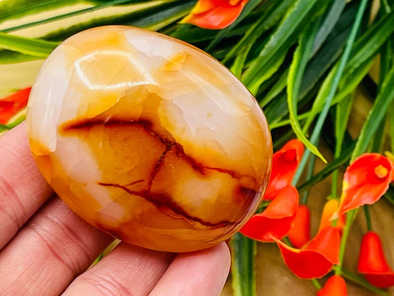 May include: A smooth, polished carnelian palm stone with a mix of orange, red, and white colors. The stone has a natural, organic shape and is perfect for holding in your hand.