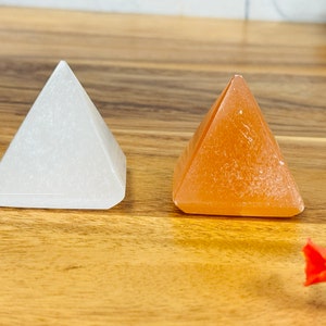 May include: Two selenite crystal pyramids, one white and one orange, on a wooden surface.