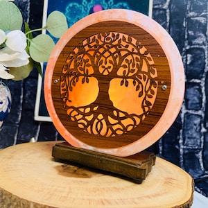 May include: A round salt lamp with a carved tree of life design. The lamp is made of Himalayan salt and has a warm orange glow. The lamp is on a wooden base.