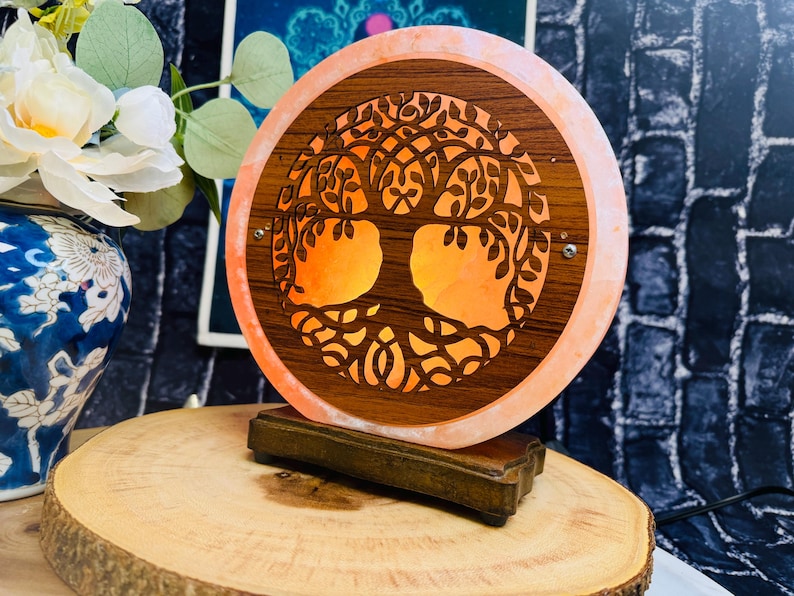 May include: A round salt lamp with a carved tree of life design. The lamp is made of Himalayan salt and has a warm orange glow. The lamp is on a wooden base.