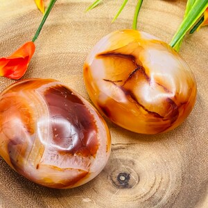 May include: Two carnelian tumbled stones, one with a white center and the other with a brown center, on a wooden surface.