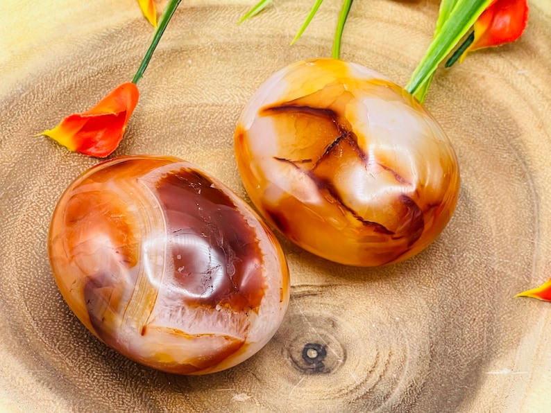 May include: Two carnelian tumbled stones, one with a white center and the other with a brown center, on a wooden surface.