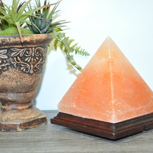 May include: A Himalayan salt pyramid lamp with a wooden base sits next to a decorative brown vase filled with greenery. The pyramid lamp is a warm orange color and is a unique home decor item.