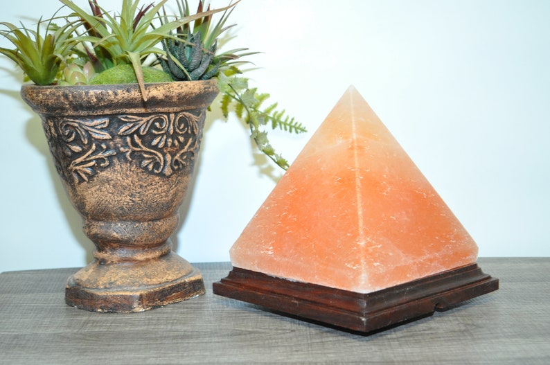 May include: A Himalayan salt pyramid lamp with a wooden base sits next to a decorative brown vase filled with greenery. The pyramid lamp is a warm orange color and is a unique home decor item.
