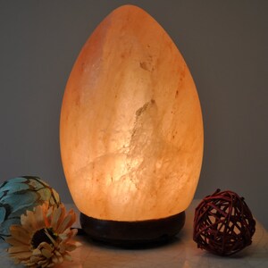 May include: A large, egg-shaped Himalayan salt lamp with a warm, orange glow. The lamp is sitting on a wooden base and is illuminated from within.