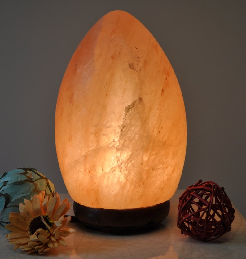 May include: A large, egg-shaped Himalayan salt lamp with a warm, orange glow. The lamp is sitting on a wooden base and is illuminated from within.