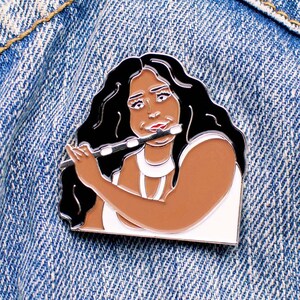 May include: A black and white enamel pin featuring a cartoon illustration of a woman playing a flute. The woman has brown skin, long black hair, and is wearing a white sleeveless top.