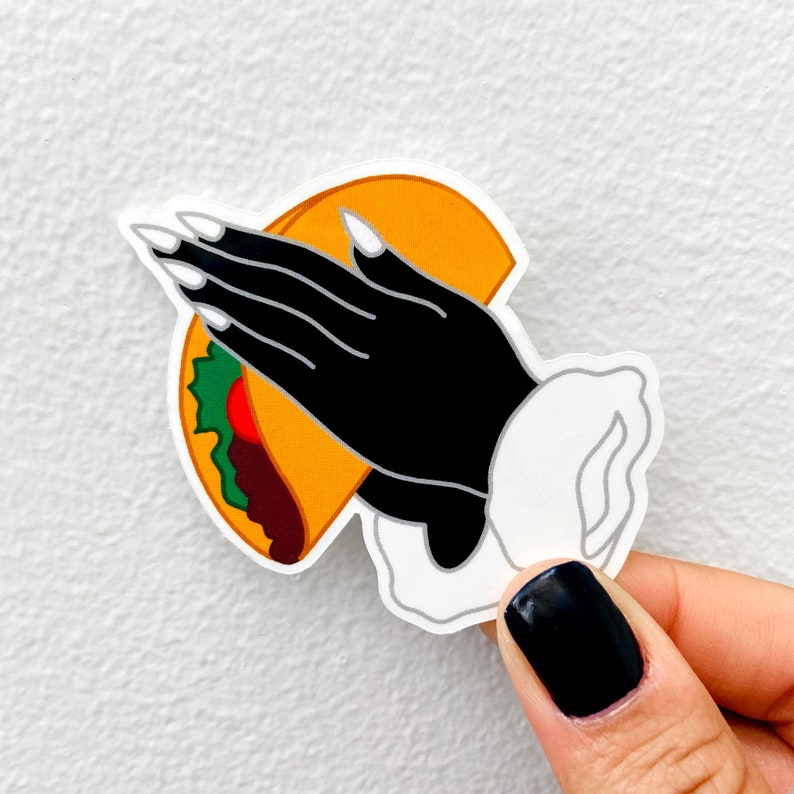 May include: A black hand with long white fingernails is holding a yellow taco with green lettuce and red meat filling. The taco is being held up in a gesture of respect or admiration.