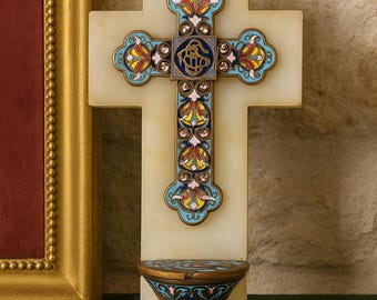 May include: A decorative cross with a detailed, colorful design. The cross is mounted on a rectangular, off-white base. The cross features blue, yellow, and red accents. A small, ornate shelf is attached below. The cross is likely a religious item.