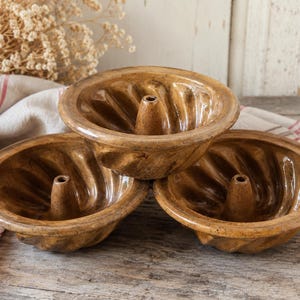 May include: Three brown ceramic bundt cake pans with a central tube and fluted sides are stacked on a wooden surface. A white and red striped cloth and dried flowers are in the background.