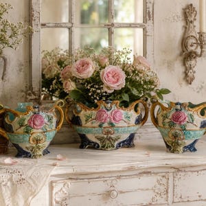 May include: Three antique ceramic vases with floral designs. The vases are arranged on a white dresser. Each vase has a light blue and gold trim, with pink roses and green leaves. The background includes a window and a vase of flowers.