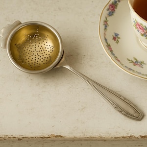 May include: A silver tea infuser with a perforated gold-colored bowl and long handle rests on a white surface. A teacup and saucer with floral designs are in the background, filled with tea. The tea cup and saucer have a gold trim.