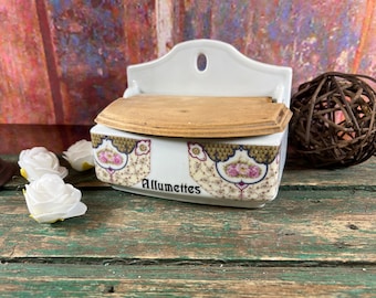 May include: White porcelain matchbox holder with a wooden lid. The front of the box has a floral design and the word "Allumettes" printed in black.