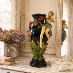 May include: A black ceramic vase with a sculpted figure and floral accents. The vase features a dark base, green and yellow leaves, and colorful flowers. A light-skinned figure is draped over the side. The vase is displayed on a white surface.