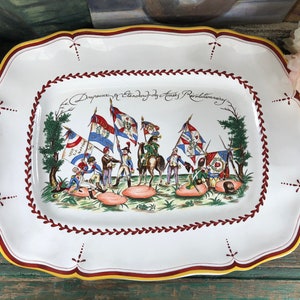 May include: A white ceramic serving platter with a red and yellow border. The platter features a hand-painted scene of soldiers marching with flags, surrounded by green foliage. The text "Depoucement et Standard des Armes Revolutionnaires" is written in black cursive.
