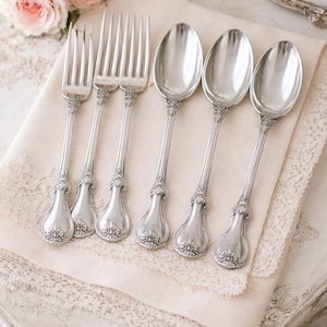 Peut inclure: Un ensemble de six fourchettes et cuillères antiques argentées. Les fourchettes ont quatre dents et les cuillères ont des cuillères ovales. Les manches présentent un motif floral orné. La coutellerie est disposée sur une serviette en lin crème.