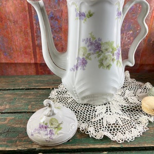 May include: White porcelain teapot with a floral design in shades of purple and green. The teapot has a lid with a floral design and a gold trim. The teapot is sitting on a white crocheted doily.