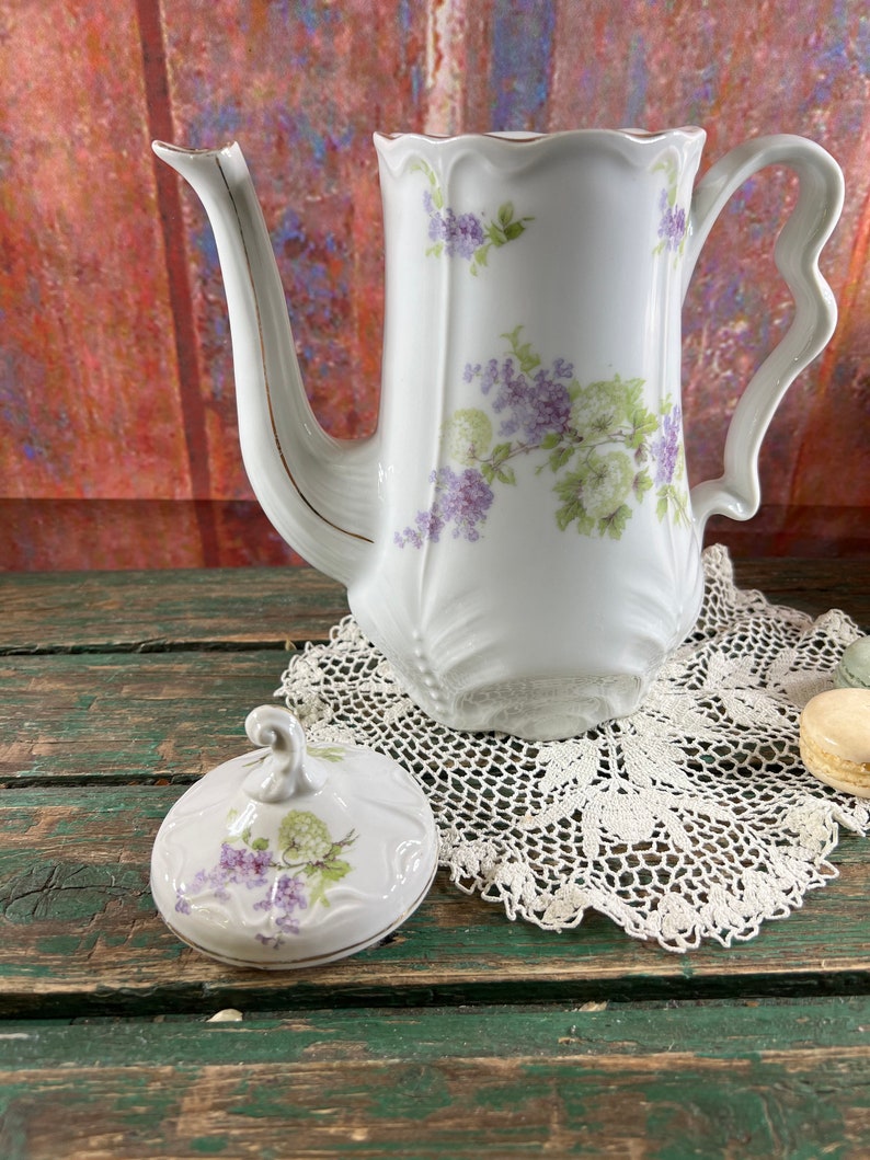 May include: White porcelain teapot with a floral design in shades of purple and green. The teapot has a lid with a floral design and a gold trim. The teapot is sitting on a white crocheted doily.