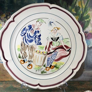 May include: A white ceramic plate with a scalloped edge and a burgundy rim. The plate features a hand-painted scene of a man and woman in traditional clothing, holding a basket of fruit. The scene is surrounded by a green and yellow floral border. The plate is signed "J. Wijffels".