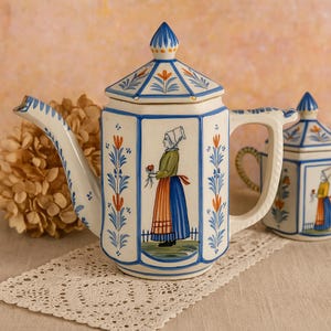 Puede incluir: Una tetera de cerámica blanca con un diseño floral azul y naranja y una ilustración de una mujer holandesa. La tetera tiene un cuerpo hexagonal, un pico curvo y un asa. Un azucarero a juego es visible en el fondo.