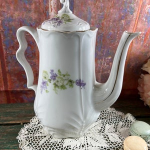 May include: White porcelain teapot with a floral design featuring purple and green flowers. The teapot has a gold trim around the lid and spout. The teapot is sitting on a white crocheted doily.