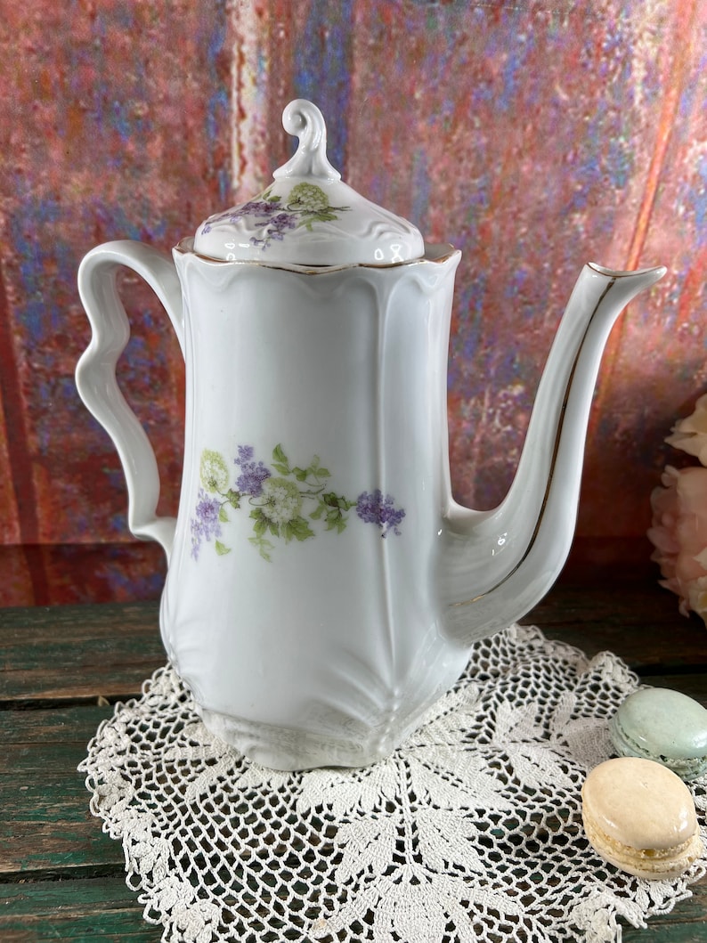 May include: White porcelain teapot with a floral design featuring purple and green flowers. The teapot has a gold trim around the lid and spout. The teapot is sitting on a white crocheted doily.