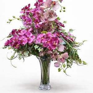 May include: A striking display of orchids in a clear glass vase. The flowers are predominantly purple and pink, with green foliage providing a contrast. The vase sits against a plain white backdrop, highlighting the floral arrangement.