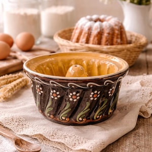 Puede incluir: Un molde para bizcocho bundt de cerámica marrón y amarilla con un diseño floral. El molde está sobre un paño de lino, con una cuchara de madera y otros ingredientes para hornear en el fondo. Un pastel horneado está en una cesta.