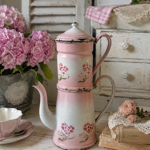 Puede incluir: Una cafetera de esmalte alta de dos niveles con un acabado degradado en rosa y blanco. La pieza está decorada con pequeños motivos florales rosas pintados a mano, incluye una tapa a juego, un pico curvo y dos asas laterales con desgaste.